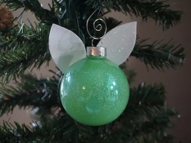 round green glitter ornament with white glitter wings on the back hanging on a evergreen tree
