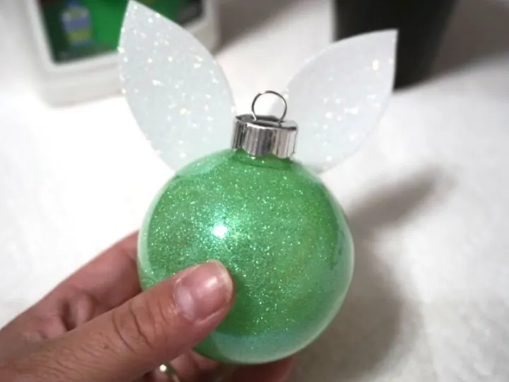  hand holding a finished tinker bell ornament, green glitter decorated ball with white glitter wings on back, paper towel background 
