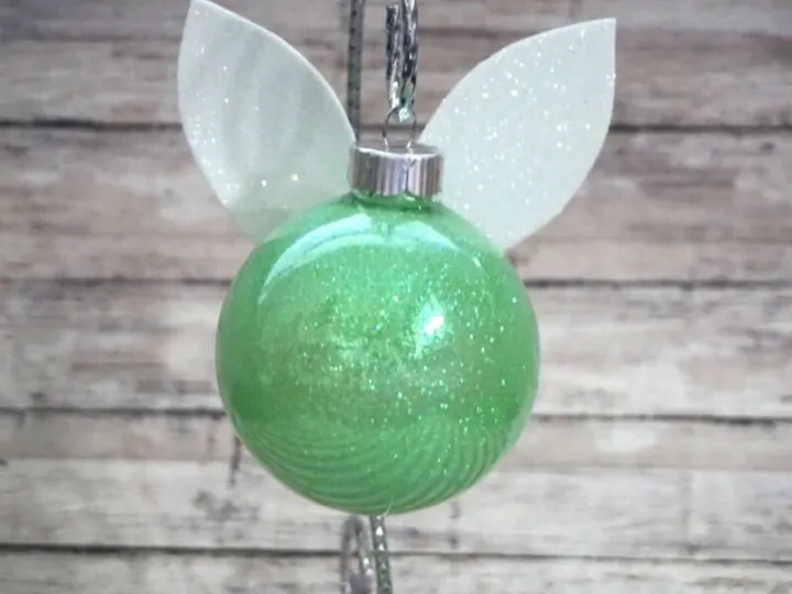 finished tinker bell ornament, green glitter decorated ball with white glitter wings on back hanging with a wood background 